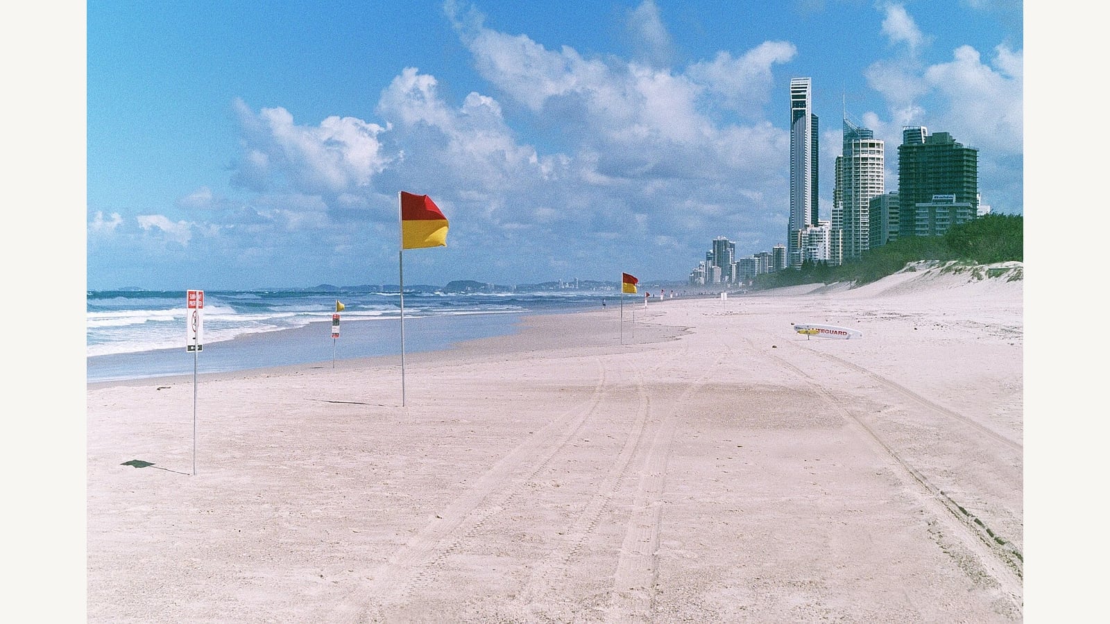 Red and yellow patrol flags on a sandy beach with surf and buildings beyond. Historical photograph, not current beach conditions.