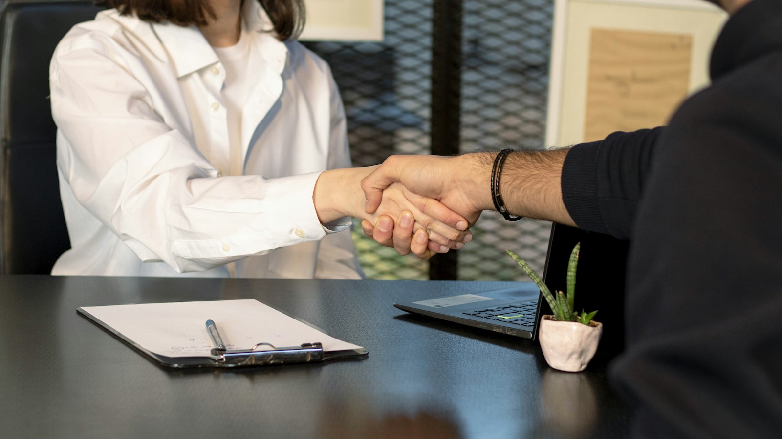 Two people shaking hands across a desk with a laptop