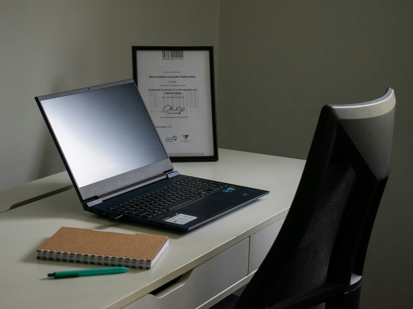 Laptop, notebook and pen on a desk beside a chair.